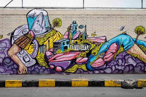 Pared de ladrillo de color blanco con la figura de un hombre con forma de ciudad. La cabeza es una especie de tiburón en un frasco. En el tronco hay edificaciones y en las piernas un árbol.