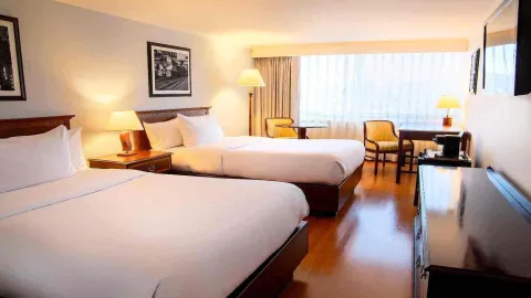 Una habitación de hotel con dos camas dobles, escritorio y una mesa pequeña con sillas. La decoración es clásica con obras de arte en blanco y negro y abundante luz natural.