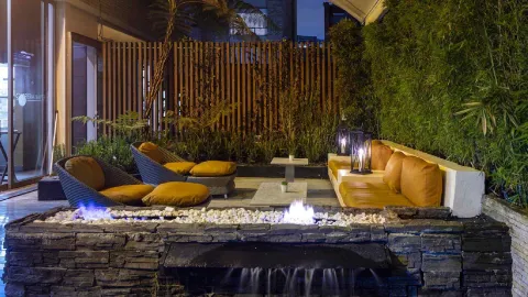 Un patio exterior elegante y acogedor con un fogón, asientos confortables con cojines naranjas y mucha vegetación, ideal para relajarse por la noche.