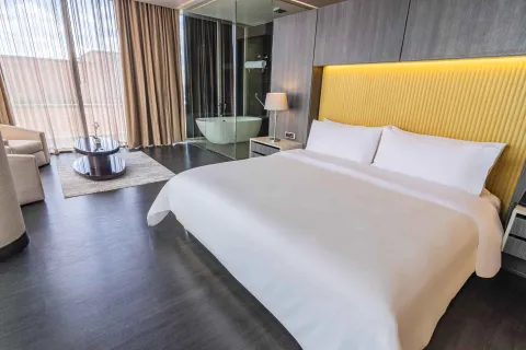 Una lujosa habitación de hotel con una cama king-size, cabecera amarilla, y un baño con bañera independiente visible a través de un panel de cristal.