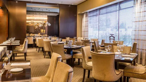 Restaurante: Restaurante moderno con sillas beige, mesas ordenadas, ventanales con persianas y lámparas. Ambiente y servicio amable.