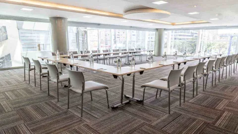  Una sala de reuniones dispuesta en forma de "U" con mesas y sillas. Grandes ventanales ofrecen vistas de la ciudad, creando un ambiente de trabajo dinámico.