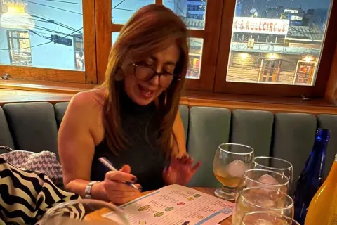  Una mujer de mediana edad con anteojos está sentada alrededor de una mesa en la que hay 4 copas y dos botellas, parece que con licor. 