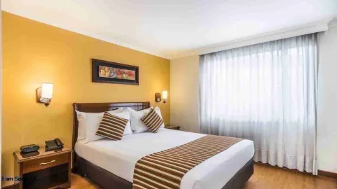 Un dormitorio de hotel con una cama doble, mesitas de noche y un televisor. La habitación es luminosa, con cortinas blancas y paredes en tonos amarillos.