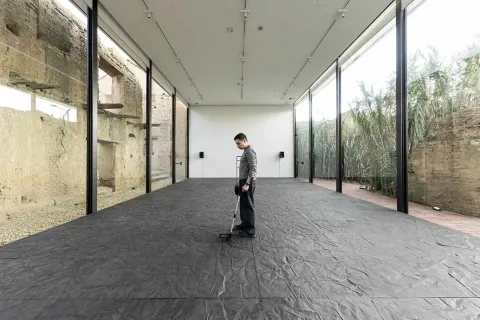 Hombre joven de pie en un salón sobre un piso, que parece ser de baldosa de color negro, mira al suelo y lo toca con un aparato que parece un detector de metáles. 