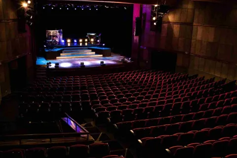 Plano general de un teatro en el que con poca iluminación se observan la silletería y la tarima, en la que hay algunas luces.