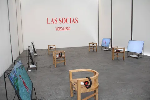 Un salón con varias pantallas de televisión sobre el suelo con una silla al frente cada una y en ellas un par de audífonos grandes. Hay una pared gris al frente con una leyenda en letras rojas que dice: “Las Socias Videojuego”.