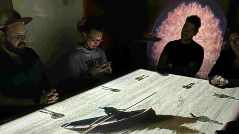 Grupo de tres hombres jóvenes a oscuras alrededor de una mesa, observan una reproducción de video en la mesa.