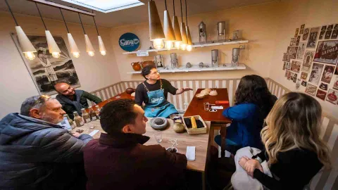 Grupo de personas jóvenes reunidas en un salón escuchando a un joven vestido con un delantal azul. En las paredes hay fotos y en la mesa varios frascos con alimentos.