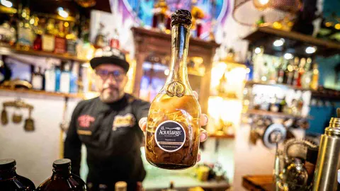  Hombre en plano borroso muestra una botella en primer plano con una etiqueta cuya leyenda dice: "El Aquelarre".