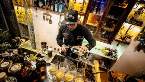 Hombre joven sirviendo algo parecido a alcohol en copas de cristal y metal.  Detrás del hombre en una pared hay muchos objetos de decoración.