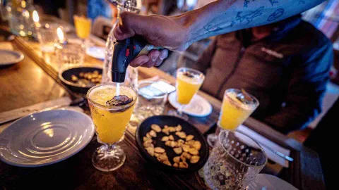 Primer plano de una mano, parece de hombre, que inyecta un líquido en varias copas de cristal. Las copas están sobre una mesa en la que también hay varios platos con trozos de alimentos.
