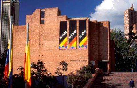 Facade of a large brick building with prominent signs in blue letters that read: “25 YEARS AT THE MAM”. A Colombian flag and a Bogotá flag are displayed to one side.