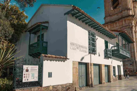 Vista de lado de la fachada de una casa antigua de estilo colonial con balcones y puertas de color verde. Resalta una leyenda en una de las paredes blancas, que dice: “Museo de la Independencia - Casa del Florero”.