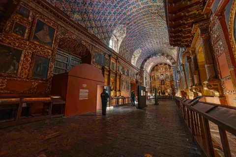 Interior de un recinto religioso, adornado por varios cuadros y retablos. El techo está adornado con unas flores azules y doradas.