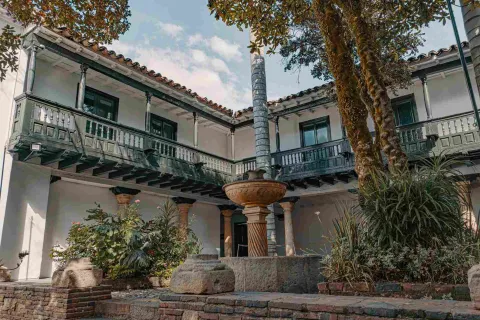Patio interior de casa antigua en la que sobresalen unos balcones en el segundo piso y una fuente de agua en el centro, rodeada por árboles y otra vegetación.