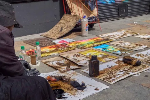 A series of paintings, seemingly freshly made, are displayed on the sidewalk. The artist can be seen from one side.
