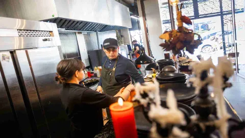 Dos chefs interactuando en una cocina profesional, con utensilios y una vela encendida en primer plano. 
