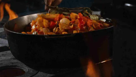 Cocción de pimientos y cebollas en una olla grande de metal sobre una llama abierta intensa.