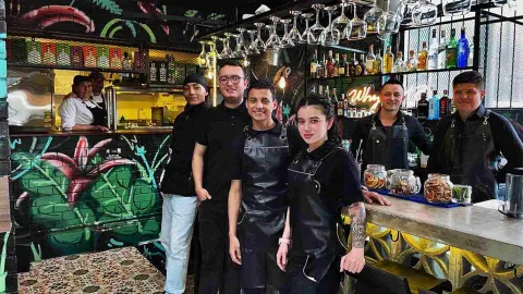 Equipo de cocina y bar sonriendo detrás de una barra moderna con botellas y copas. 