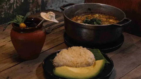  Bodegón con una olla de comida, un plato de arroz con aguacate y una bebida en un recipiente de barro sobre una mesa.