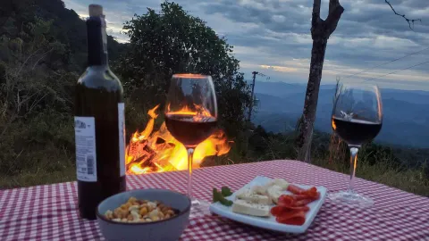 Botella de vino, dos copas, frutos secos y una tabla de quesos sobre un mantel a cuadros, con una fogata y vistas a la montaña al anochecer.