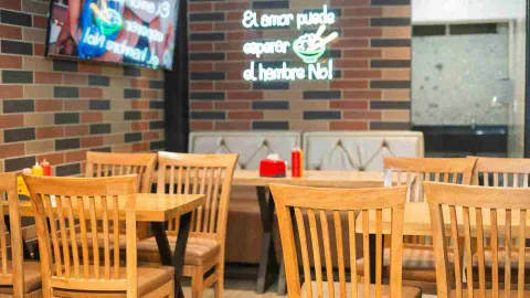 Interior del restaurante con mesas y sillas de madera, y un letrero de neón en una pared de ladrillo que dice "El amor puede esperar, el hambre No!".