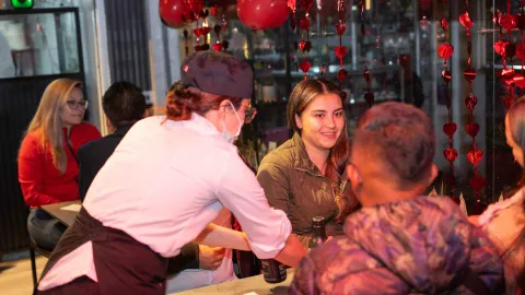 Camarera con mascarilla y gorra sirve a una cliente sonriente en un restaurante decorado con corazones rojos.