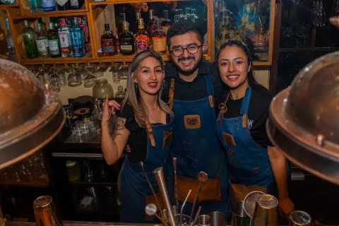 Tres sonrientes bartenders, dos mujeres y un hombre, posan detrás de una barra bien surtida. 