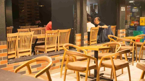 Dos hombres conversan sentados en una mesa de madera dentro del restaurante, con otros clientes visibles al fondo.