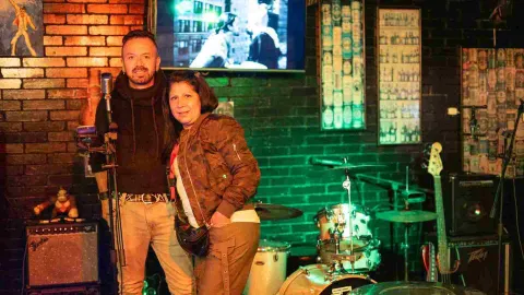 Una pareja posa en el escenario del bar, con una batería y un bajo detrás de ellos, bajo luz verde y ámbar.