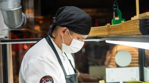 Chef de sushi con uniforme, gorro negro y mascarilla blanca, enfocado en la preparación de alimentos.