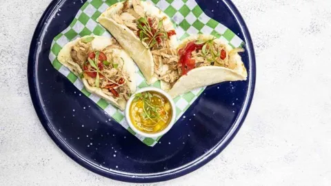 Tres tacos de tortilla de maíz rellenos de pollo deshebrado, aderezos y acompañados de una salsa. 
