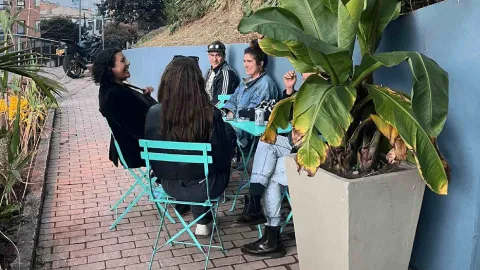 Cuatro personas conversan sentadas alrededor de una mesa turquesa en la terraza, junto a una maceta grande.