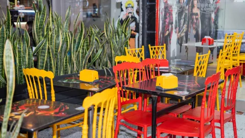 Área de comedor al aire libre con mesas temáticas de cómics, sillas rojas y amarillas, y una fila de plantas Sansevieria.