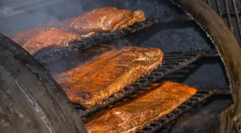 Costillas de cerdo asándose lentamente en una parrilla humeante, mostrando su delicioso ahumado. 