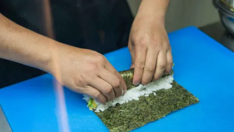 Manos de una persona enrollando un rollo de sushi con arroz y aguacate sobre una tabla de cortar azul.