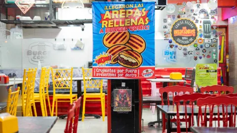 Un gran cartel azul anuncia "Arepas Rellenas" en el interior del restaurante Burger Comic, decorado con pegatinas.