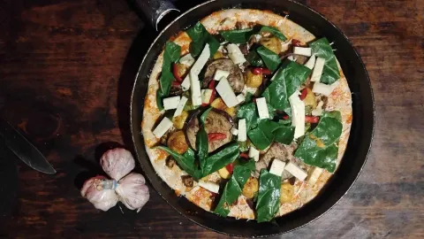 Vista cenital de una pizza con espinacas, berenjena y queso, cocinada en una sartén negra, con una cabeza de ajo al lado sobre la mesa.