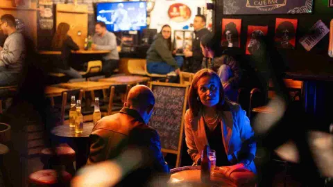 Dos personas conversan en primer plano en una mesa del bar, con otros clientes al fondo.