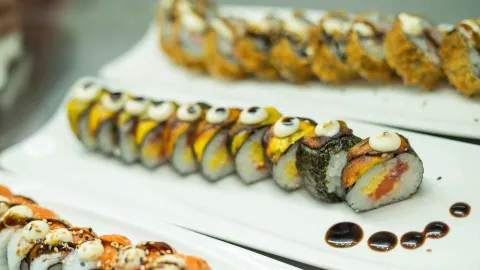  Plato de sushi con una variedad de rollos, algunos con plátano frito y salsas, presentados en una fila.