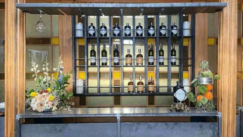 Barra de bar con estantería de metal industrial que alberga botellas de ginebra y dispensadores de vidrio con cítricos frescos.