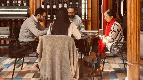 Tres personas reunidas alrededor de una mesa de madera en un restaurante con suelo de baldosas, revisando documentos y conversando.