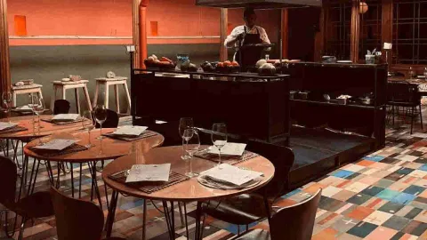 Interior de restaurante con mesas redondas de madera, sillas, suelo de baldosas de colores y un chef trabajando en la cocina abierta.