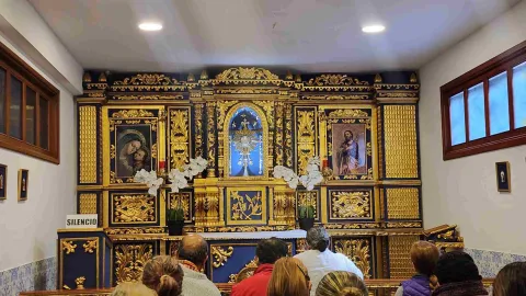  Interior de iglesia donde hay personas, parece que orando, al frente de un retablo donde hay dos cuadros con figuras de santos y la figura del denominado Santísimo. Se destacan los ricos detalles dorados de la ornamentación.