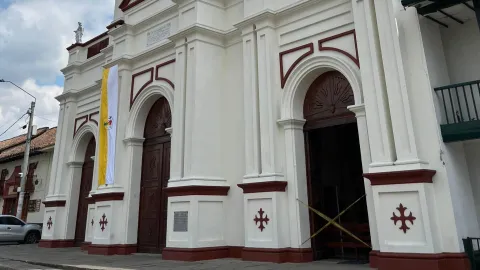 Primer plano de la parte baja de la parroquia en la que se observan tres puertas grandes en forma de arco. En la pared está colgada una bandera con dos franjas de color, una amarilla y una blanca.