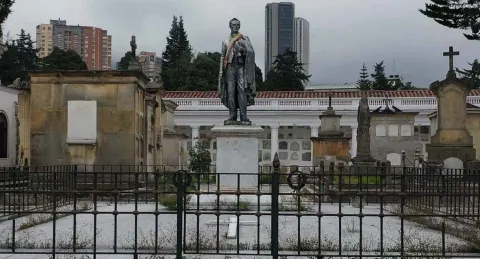 Stone statue in a mausoleum, seemingly of General Santander, with a ribbon in the colors of the Colombian flag. Several other stone mausoleums are nearby. Buildings can be seen in the distance, above the mausoleum.