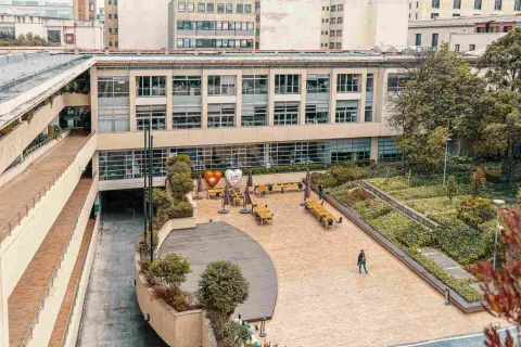  Plano general visto desde arriba del interior del palacio. Se observa una amplia plazoleta en ladrillo, rodeada de árboles. Una persona camina por entre la plazoleta. Hay una decoración con figuras de corazones y un grupo de mesas con sillas y parasoles.