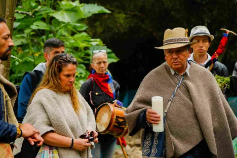 Un grupo de personas, algunas de las cuales parecen estar vestidas con atuendos tradicionales. En primer plano, un hombre lleva un sombrero de paja tejido y un poncho marrón; está sosteniendo una vela blanca. A su izquierda, una mujer con un suéter color crema está mirando un objeto pequeño en sus manos. Detrás de ella, una mujer sostiene un tambor, y lleva un pañuelo rojo, azul y amarillo alrededor del cuello. En segundo plano, se ven otras personas y vegetación. La escena parece tener lugar al aire libre.
