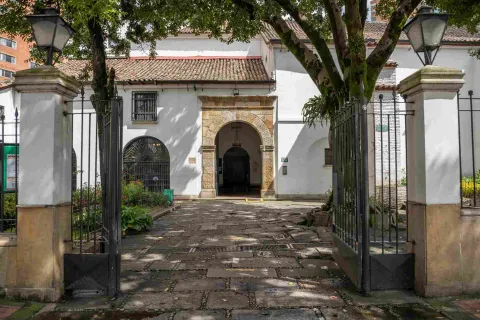  Parte exterior de la puerta principal de la iglesia en la que se ve una reja que da paso al antejardín y el arco en piedra de la puerta al fondo.